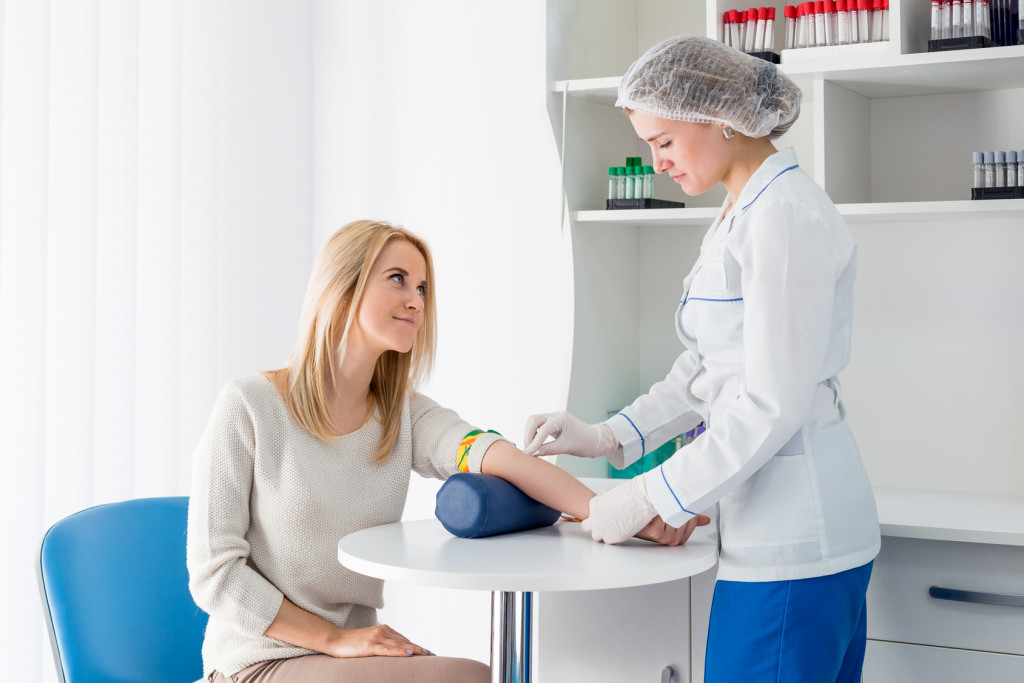 Preparation for blood test with beautiful young blond woman by female doctor in white coat medical uniform on the table in white bright room. Nurse rubbing a hand styryl patient tissue.